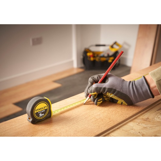 Photo of STANLEY 0-30-687 tape measure on wood, worker with gloves marking with red pencil, tool bag in background.