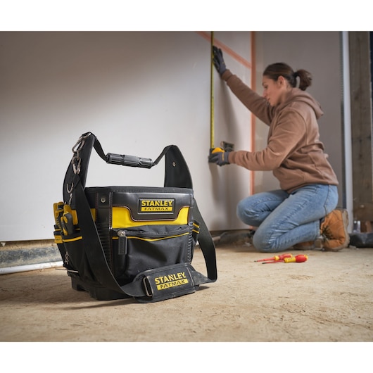 STANLEY 1-93-952 tool bag with yellow and black design on floor, worker measuring wall in background.