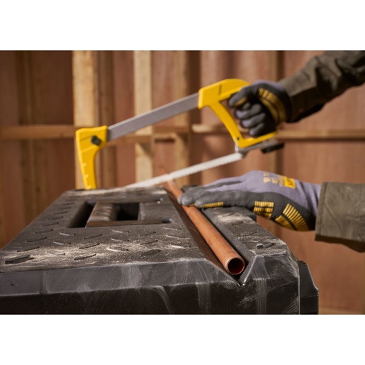 STANLEY 1-95-829 hacksaw held by gloved hands, cutting copper pipe on a black workbench, side angle.