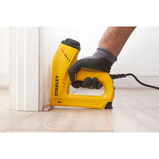 Photo of STANLEY 6-TRE550 electric stapler in use near baseboard, yellow body, side angle view.