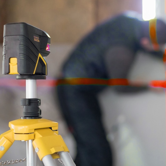 STANLEY FMHT1-77414 laser level on tripod, emitting red lines, indoor workspace, angled side view.