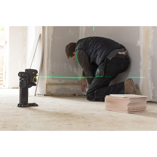 STANLEY FMHT1-77442 laser level projecting green lines, shown on a construction site floor near a worker and tiles.