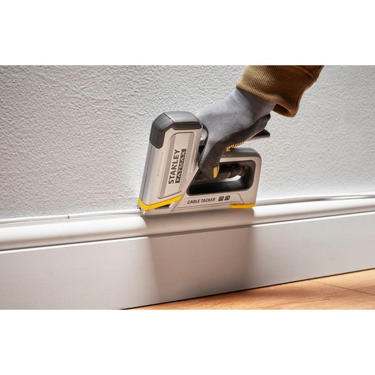 Stanley FMHT70100-0 cable tacker in use along baseboard, shown held by a gloved hand against wall.