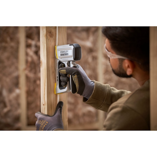 STANLEY FMHT70100-0 stud finder held by gloved hand against wooden frame in construction setting.
