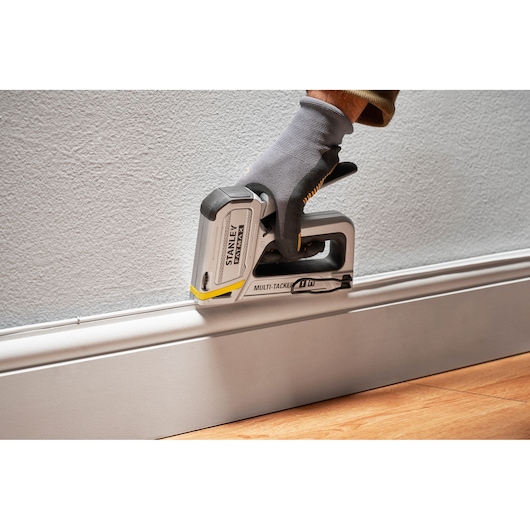 Photo of STANLEY FMHT70250-0 multi-tacker stapler in use, angled against wall trim, gloved hand holding tool.
