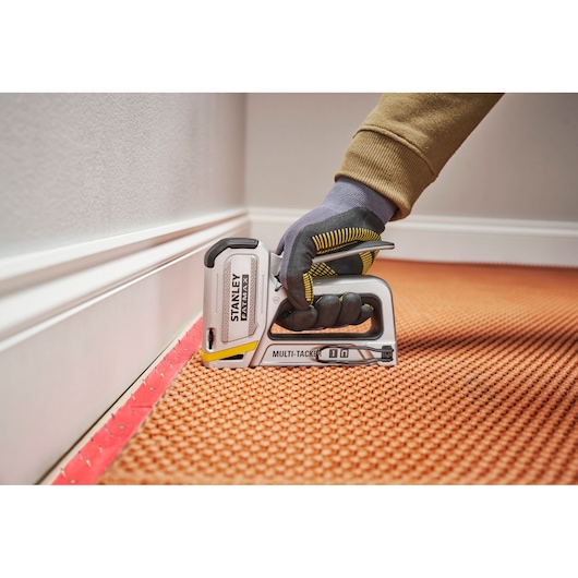 STANLEY FMHT70250-0 multi-tacker stapler in use on carpet by gloved hand near baseboard, close-up photo.