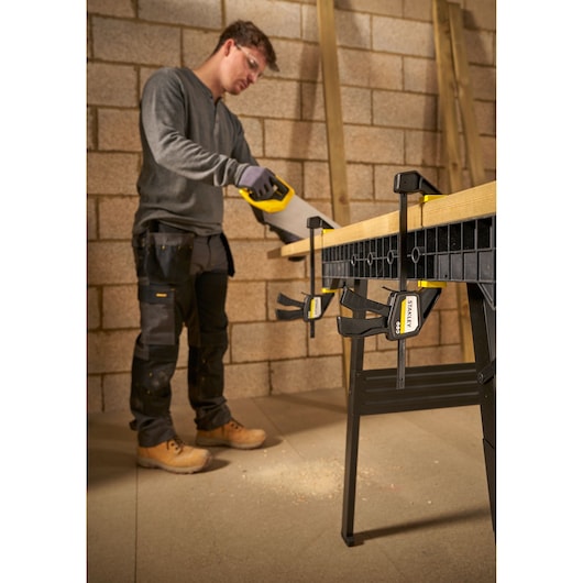 Photo of STANLEY FMHT83271-0 clamp securing wood to workbench in a workshop, worker sawing near clamps.