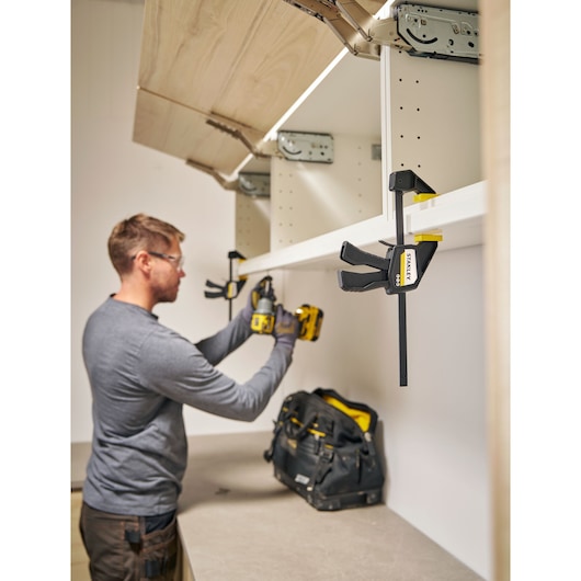 Photo of STANLEY FMHT83271-0 clamp holding a shelf with tool bag and person assembling cabinet.