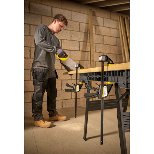 Photo of STANLEY FMHT83275-0 bar clamps securing wood to a workbench during sawing in a workshop.
