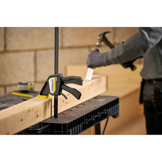 STANLEY FMHT83275-0 bar clamp securing wood beam on workbench, person using hammer in background.