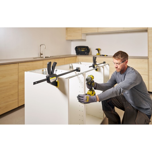 Photo of STANLEY FMHT83275-0 bar clamp holding white cabinet during assembly in kitchen, with worker using power drill.