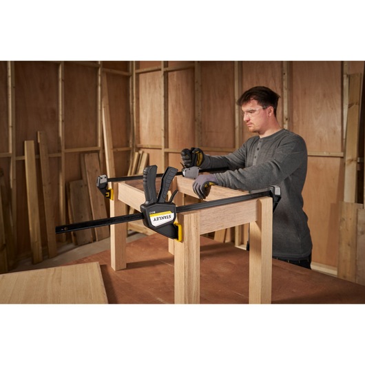 STANLEY FMHT83276-0 bar clamp securing wood frame in workshop, person assembling furniture.