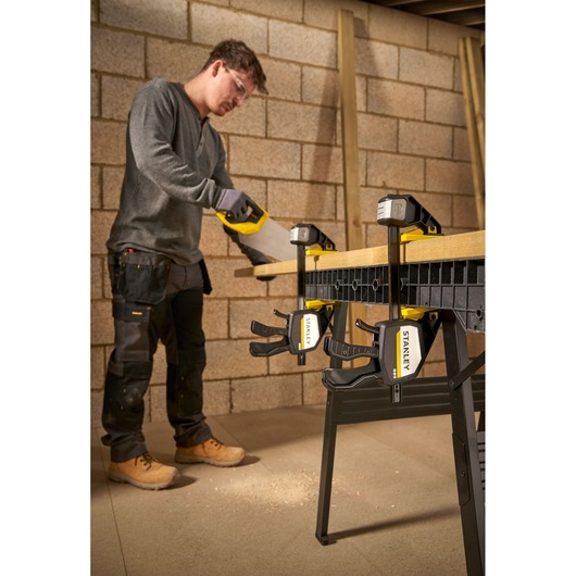 Photo of STANLEY FMHT83278-0 clamps holding wood on a workbench while a person saws, workshop background.