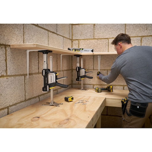 STANLEY FMHT83554-1 clamp holding wood shelf, workshop scene with tools and man assembling, front angle.