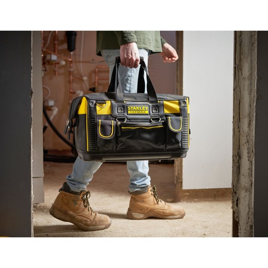 STANLEY FMST1-71180 tool bag, black and yellow, carried by person in work boots, indoor setting.