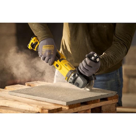 STANLEY SFMCG300B angle grinder in use cutting a concrete slab, close-up of hands wearing gloves.