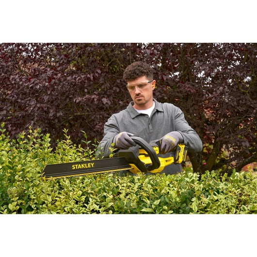 Photo of STANLEY STCHT8255M1 hedge trimmer in use, yellow and black, trimming green hedges outdoors.