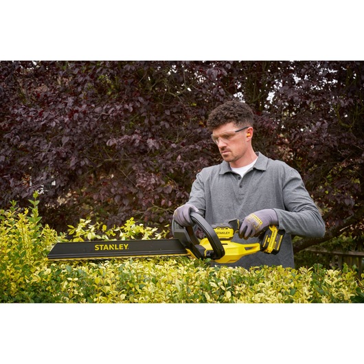 Photo of STANLEY STCHT8255M1 cordless hedge trimmer in use, yellow and black design, trimming green bushes outdoors.