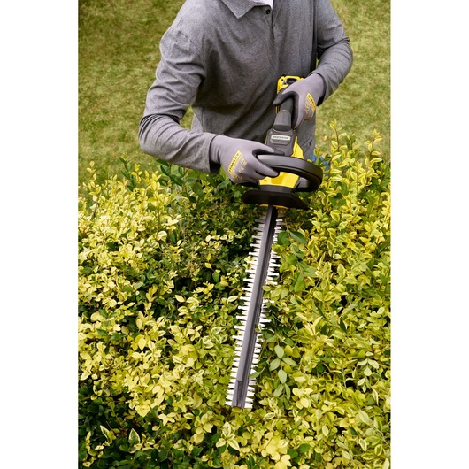 STANLEY STCHT8255M1 hedge trimmer in use, person wearing gloves trimming green bushes outdoors.