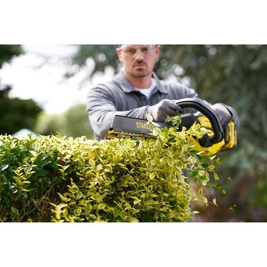 Photo of STANLEY STCHT8255M1 hedge trimmer in use, cutting a green hedge outdoors.