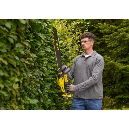 STANLEY STCHT8255M1 cordless hedge trimmer held by person trimming green hedge outdoors.