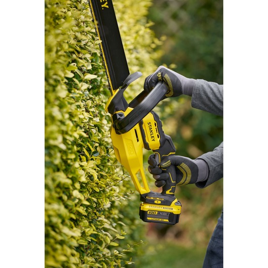 STANLEY STCHT8255M1 hedge trimmer in use, yellow and black, trimming green hedge outdoors.
