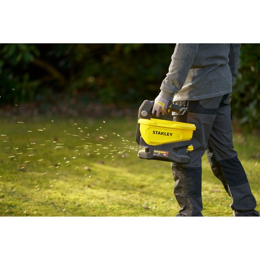Photo of a person using STANLEY STCOE300D1 handheld yellow spreader to distribute material on grass outdoors.