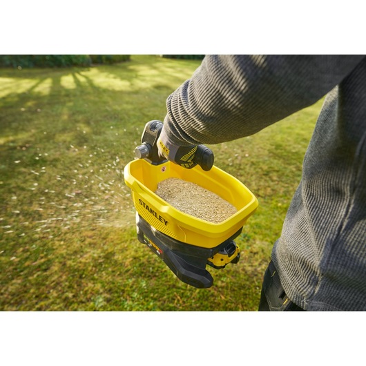 STANLEY STCOE300D1 handheld yellow broadcast spreader in use on grass, dispensing seed from above view.