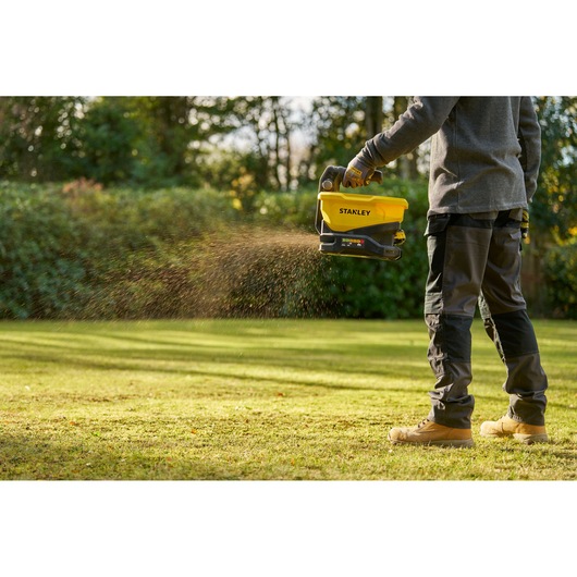 Outdoor photo of person using STANLEY STCOE300D1 handheld spreader, spreading material on grass lawn.