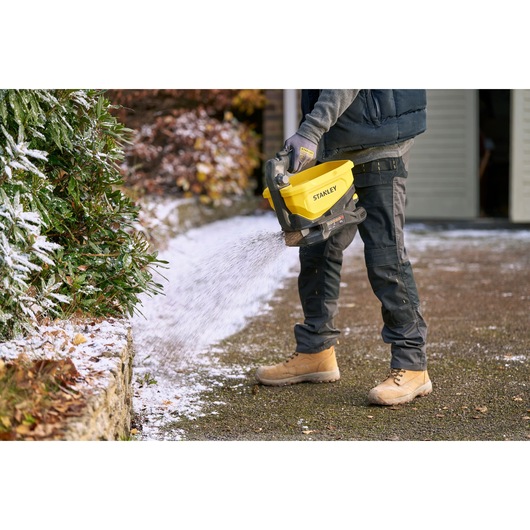 Photo of STANLEY STCOE300D1 handheld spreader in use on snowy path, yellow and black design, outdoor winter setting.