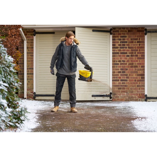 STANLEY STCOE300D1 handheld spreader being used to spread grit on a snowy driveway in front of a garage.