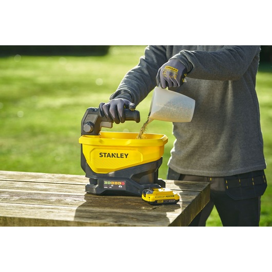 STANLEY STCOE300D1 yellow cordless spreader on wooden table, person filling with seed outdoors.