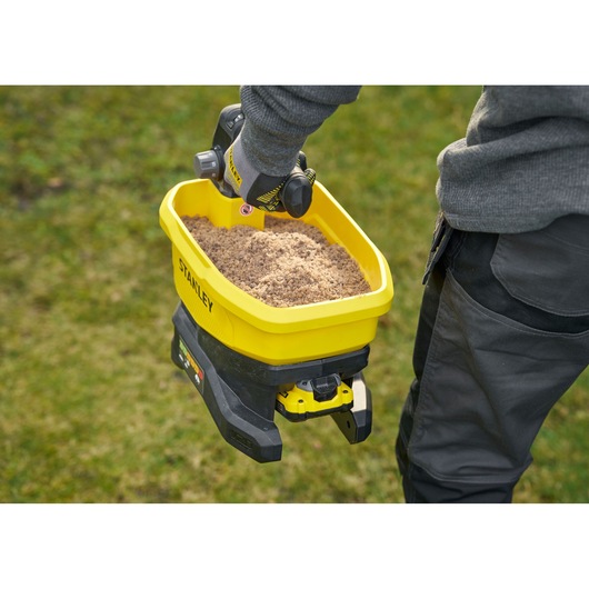 Stanley STCOE300D1 yellow spreader held by worker, filled with sand, used outdoors on grass.