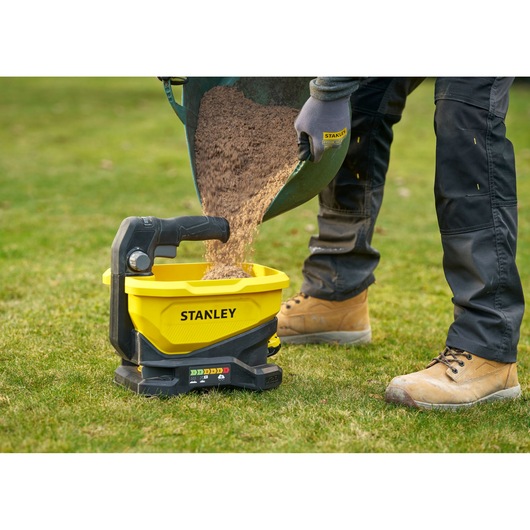 STANLEY STCOE300D1 yellow battery-powered spreader on grass, sand being poured into hopper, side angle.