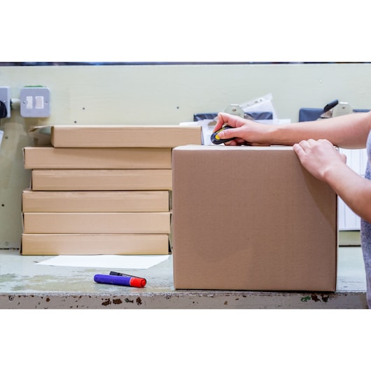 STANLEY STHT0-10292 utility knife cutting a cardboard box, stacks of boxes in background, desktop view.