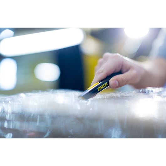 Photo of STANLEY STHT0-10293 utility knife cutting plastic wrap on a package in a warehouse setting.