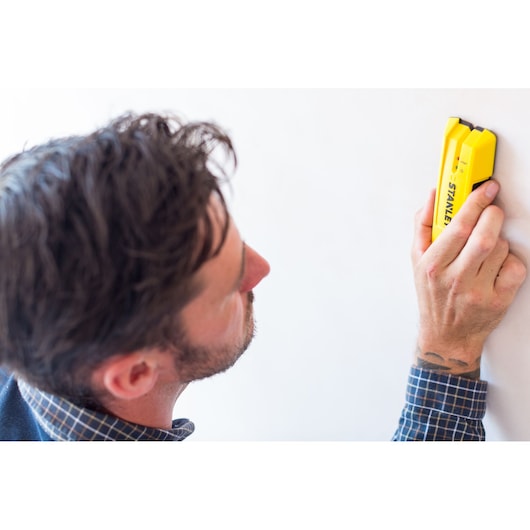Photo of STANLEY STHT0-77050 stud finder being used on a white wall, yellow handheld tool visible.
