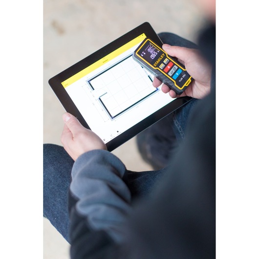 Photo of STANLEY STHT1-77343 laser distance measurer in hand, used with a tablet showing measurement data.