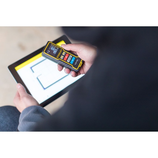 STANLEY STHT1-77343 laser distance measurer held in hand, tablet showing floor plan in background.