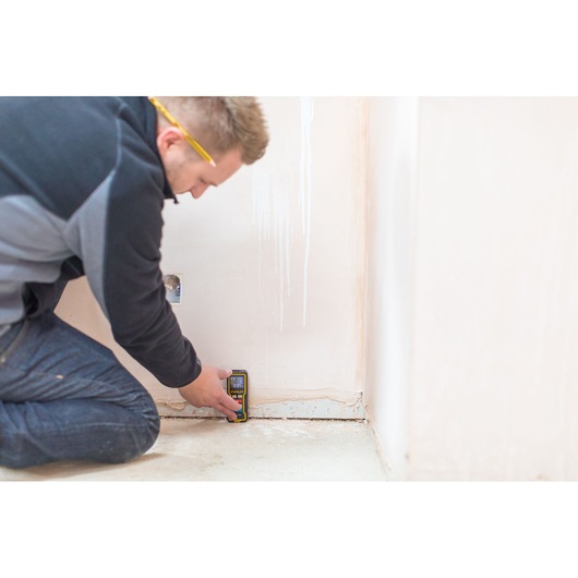 Person using STANLEY STHT1-77343 laser distance measurer near a wall, close-up indoor scene.