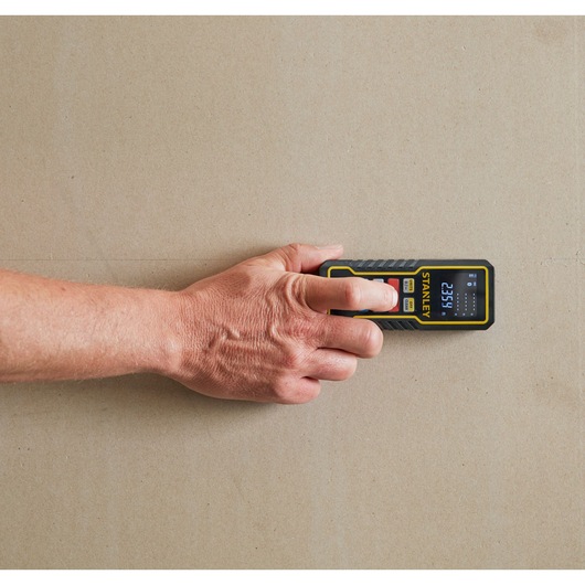 Photo of STANLEY STHT1-77343 digital distance meter in use, close-up from above, handheld against beige wall.