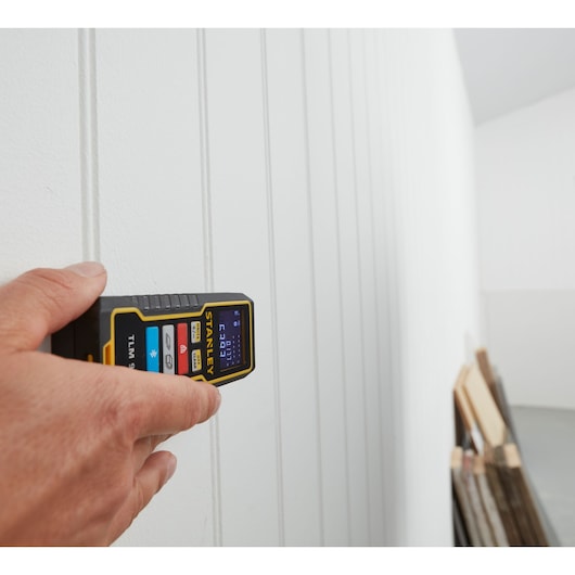 STANLEY STHT1-77343 laser distance measurer held against white paneled wall, close-up photography.