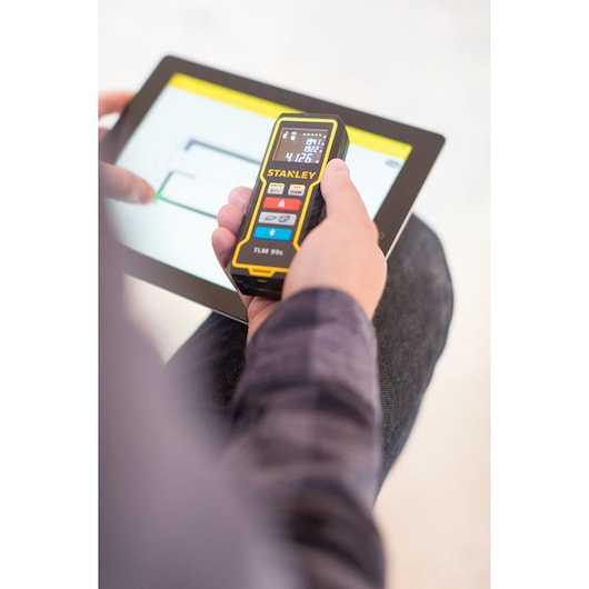 Photo of STANLEY STHT1-77343 laser distance measurer in use next to a tablet, product held in hand.