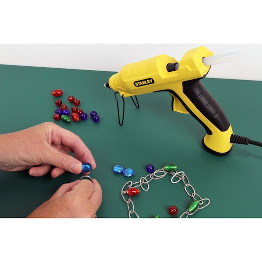 Stanley STHT6-70417 yellow glue gun for crafts shown with beads and jewelry chain on green surface.
