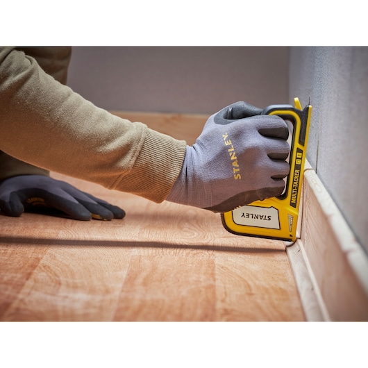 Stanley STHT70440-0 manual stapler in use, yellow and black, held by gloved hand against baseboard.